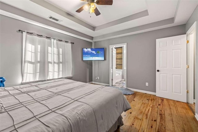 bedroom featuring light hardwood / wood-style flooring, ensuite bath, ceiling fan, and a tray ceiling