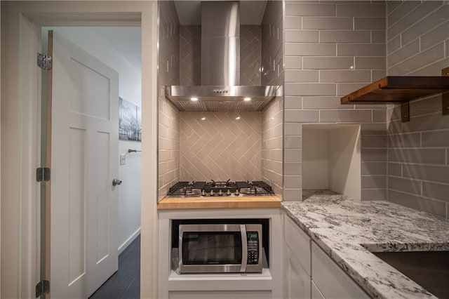 kitchen with wall chimney exhaust hood, light stone counters, appliances with stainless steel finishes, decorative backsplash, and white cabinets