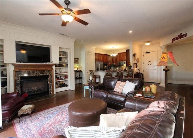 living room with crown molding, built in features, dark hardwood / wood-style floors, a high end fireplace, and ceiling fan with notable chandelier