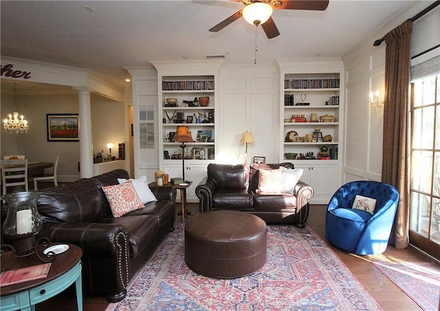 living room with crown molding, built in features, hardwood / wood-style floors, ceiling fan with notable chandelier, and ornate columns