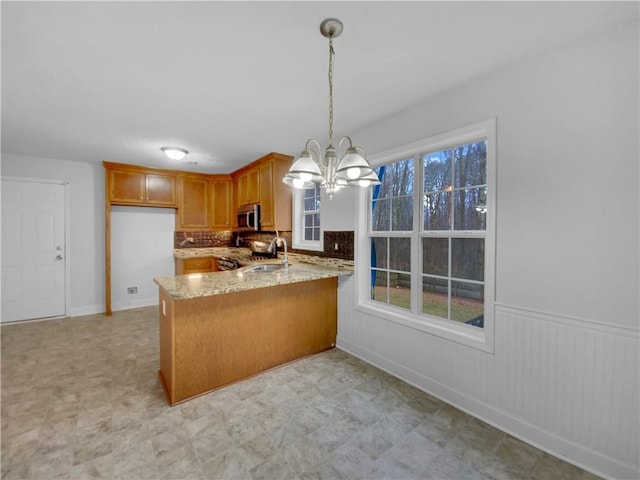 kitchen featuring brown cabinetry, stainless steel microwave, light stone counters, decorative light fixtures, and a peninsula