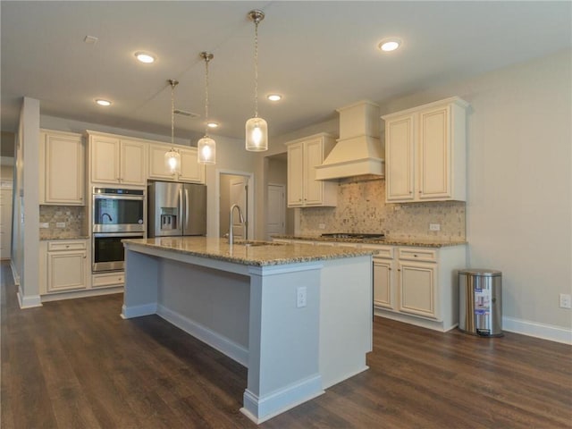 kitchen with premium range hood, decorative light fixtures, sink, stainless steel appliances, and a center island with sink