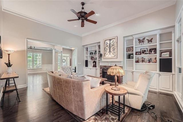 living area featuring built in shelves, a ceiling fan, baseboards, dark wood-style flooring, and crown molding