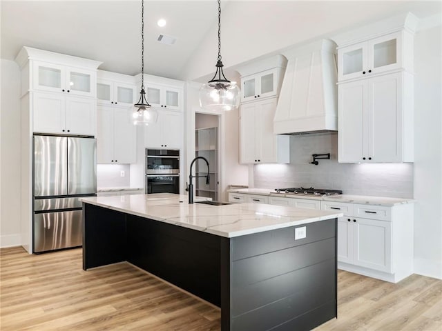 kitchen with a center island with sink, custom range hood, white cabinetry, appliances with stainless steel finishes, and decorative light fixtures