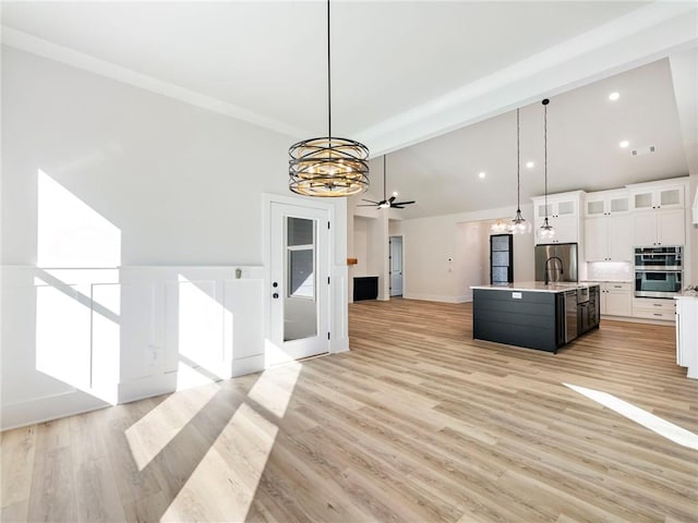 kitchen with light wood-type flooring, appliances with stainless steel finishes, decorative light fixtures, an island with sink, and white cabinets