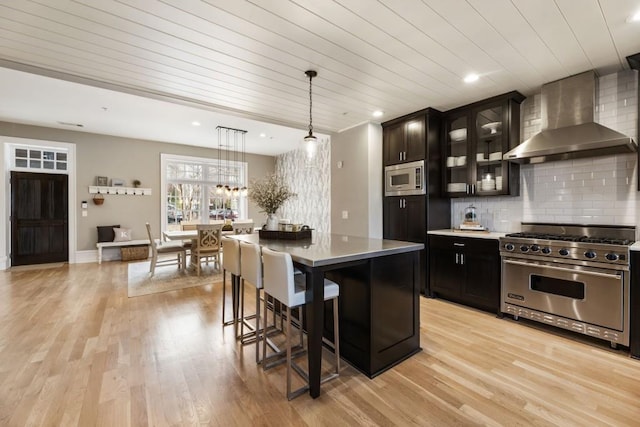 kitchen with light wood finished floors, stainless steel appliances, decorative backsplash, glass insert cabinets, and wall chimney exhaust hood