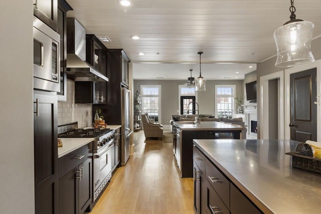 kitchen featuring wall chimney range hood, open floor plan, appliances with stainless steel finishes, a fireplace, and a sink