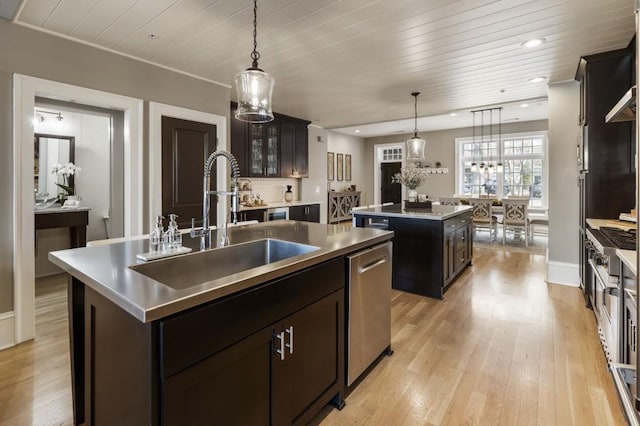 kitchen featuring a kitchen island with sink, a sink, light wood-style floors, stainless steel counters, and stainless steel dishwasher