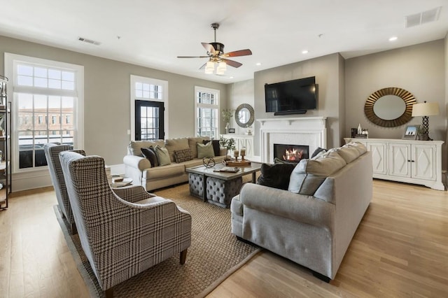 living room with a lit fireplace, visible vents, light wood-style flooring, and a ceiling fan