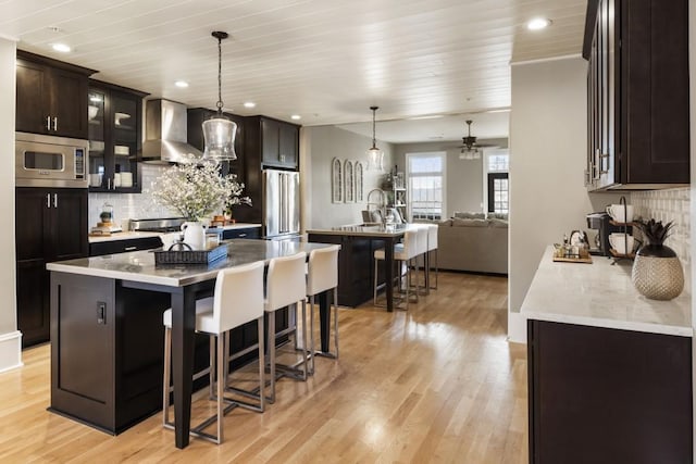 kitchen featuring a kitchen island, a kitchen bar, appliances with stainless steel finishes, light wood-style floors, and wall chimney exhaust hood