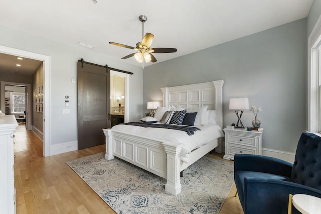 bedroom with baseboards, light wood-type flooring, a barn door, ensuite bath, and a ceiling fan