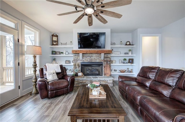 living area with a stone fireplace, built in shelves, ceiling fan, and wood finished floors