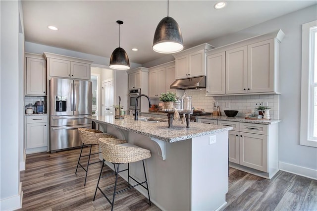 kitchen with under cabinet range hood, an island with sink, appliances with stainless steel finishes, wood finished floors, and a sink