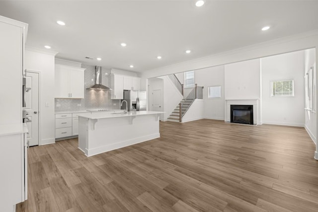 kitchen with white cabinetry, a kitchen breakfast bar, a kitchen island with sink, stainless steel fridge with ice dispenser, and wall chimney range hood