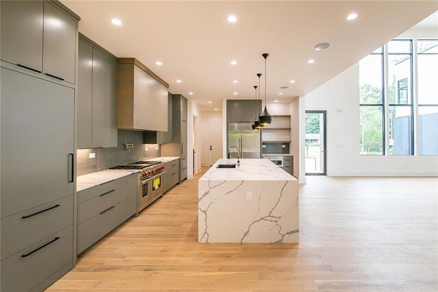 kitchen with backsplash, light hardwood / wood-style flooring, hanging light fixtures, premium appliances, and an island with sink