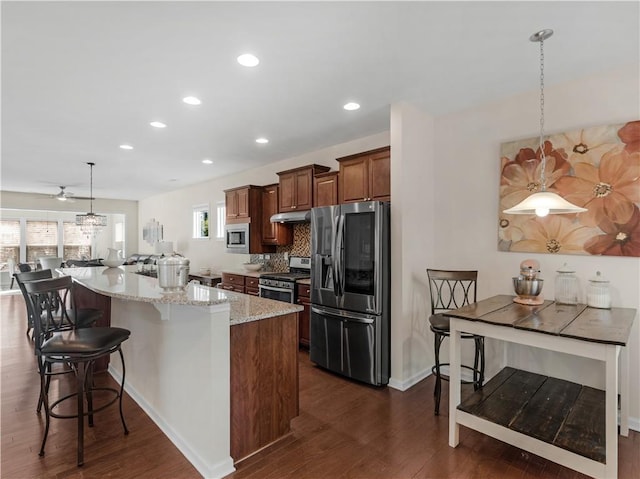 kitchen featuring dark wood-style flooring, under cabinet range hood, appliances with stainless steel finishes, a kitchen bar, and tasteful backsplash