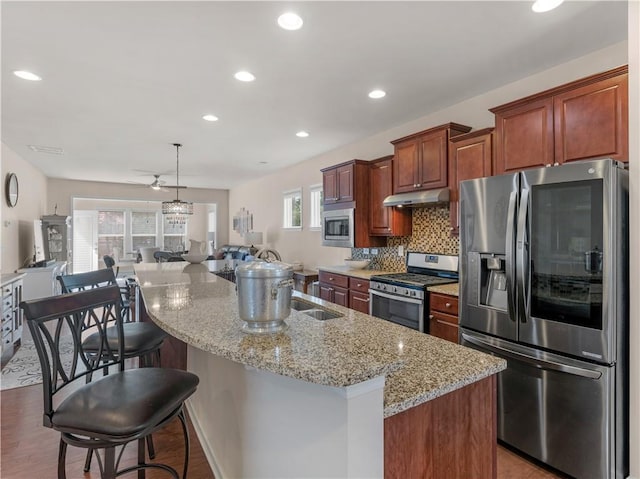 kitchen with an island with sink, stainless steel appliances, decorative backsplash, under cabinet range hood, and open floor plan