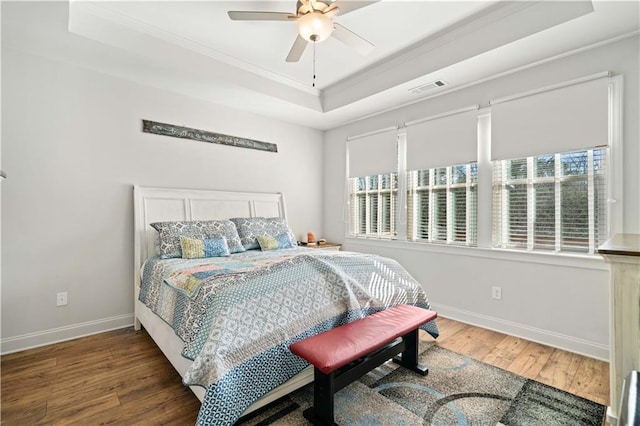 bedroom featuring wood finished floors, visible vents, baseboards, a tray ceiling, and crown molding