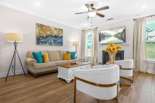 living room with light wood-style flooring, a glass covered fireplace, visible vents, ceiling fan, and crown molding