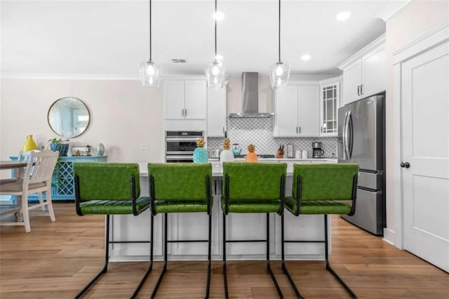 kitchen featuring backsplash, a kitchen bar, white cabinets, wall chimney exhaust hood, and appliances with stainless steel finishes