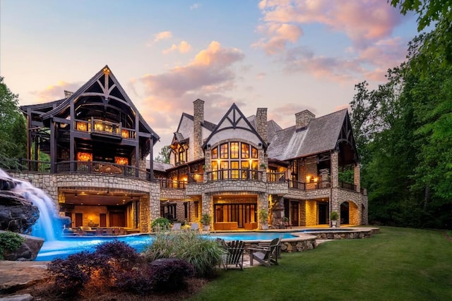 back of property at dusk featuring stone siding, an outdoor pool, a chimney, a balcony, and a lawn