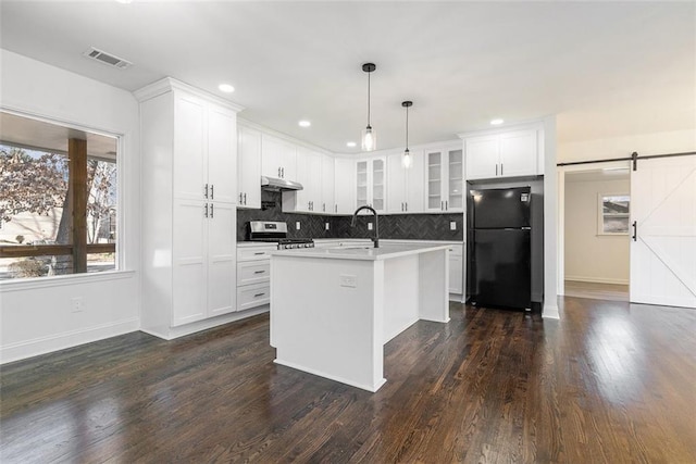 kitchen with a kitchen island with sink, black fridge, hanging light fixtures, stainless steel stove, and a barn door