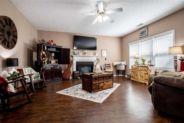 living area featuring dark wood-style floors, visible vents, a ceiling fan, and a glass covered fireplace