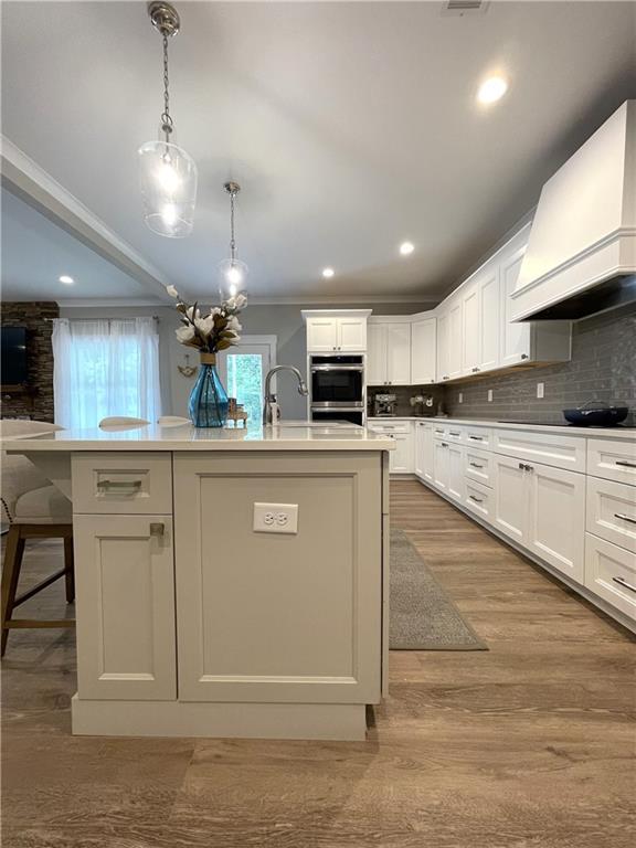 kitchen featuring premium range hood, decorative light fixtures, a kitchen island with sink, backsplash, and white cabinetry