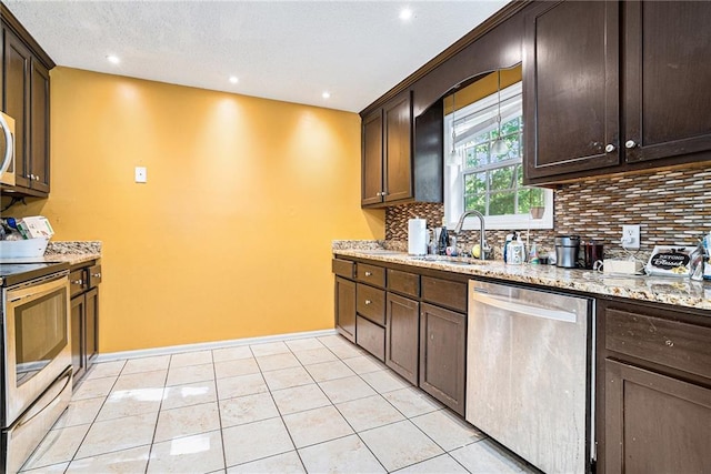 kitchen featuring appliances with stainless steel finishes, a sink, tasteful backsplash, dark brown cabinets, and light stone counters