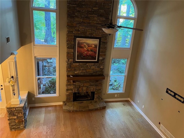 unfurnished living room with ceiling fan, a towering ceiling, a stone fireplace, and hardwood / wood-style flooring