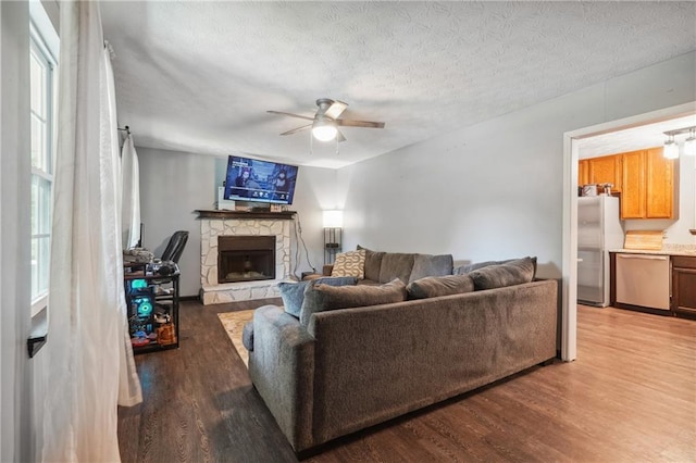 living room featuring wood-type flooring, a textured ceiling, a stone fireplace, and ceiling fan