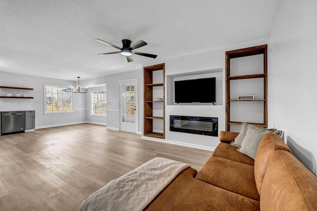 living room featuring built in features, baseboards, a glass covered fireplace, wood finished floors, and ceiling fan with notable chandelier