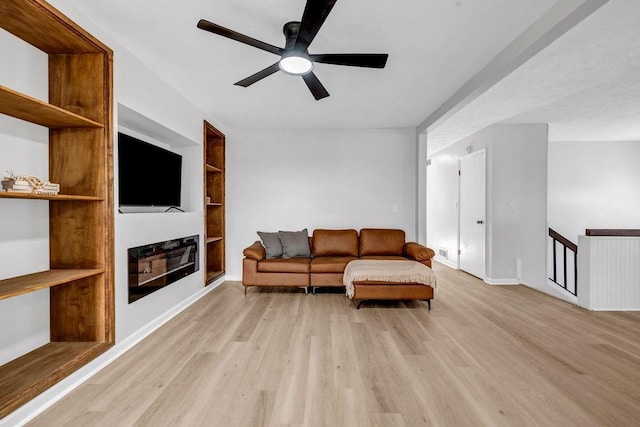 living area featuring built in shelves, a glass covered fireplace, light wood-style flooring, and a ceiling fan