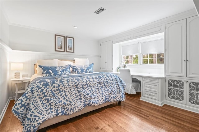 bedroom featuring built in study area, ornamental molding, a wainscoted wall, visible vents, and wood finished floors