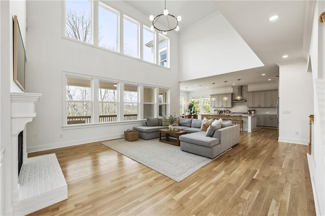 living room featuring a fireplace with raised hearth, baseboards, a chandelier, light wood-style floors, and a high ceiling