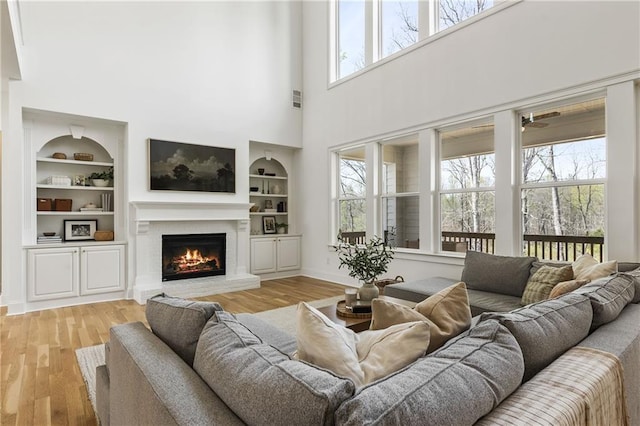 living area featuring light wood finished floors, visible vents, built in shelves, a lit fireplace, and a towering ceiling
