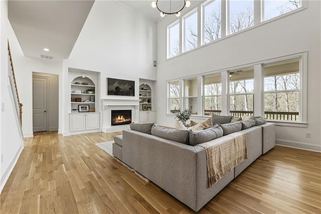 living room with a warm lit fireplace, built in features, a high ceiling, baseboards, and light wood-style floors