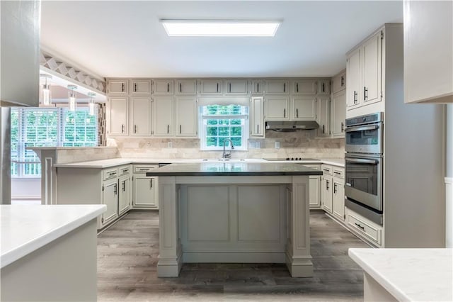 kitchen with a sink, stainless steel double oven, under cabinet range hood, backsplash, and wood finished floors