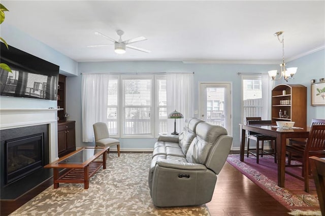 living room with wood finished floors, baseboards, a glass covered fireplace, crown molding, and ceiling fan with notable chandelier