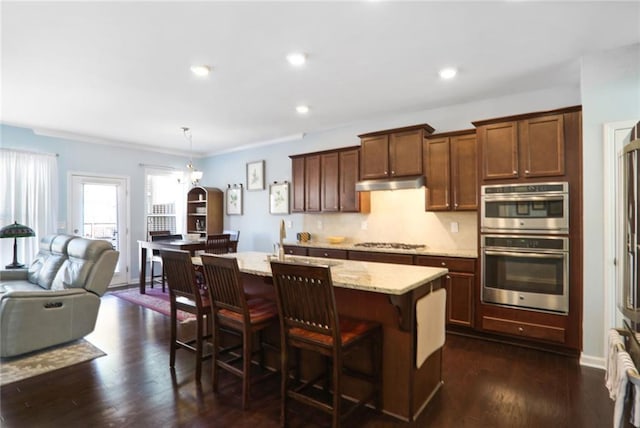 kitchen featuring under cabinet range hood, light stone counters, open floor plan, appliances with stainless steel finishes, and dark wood-style flooring