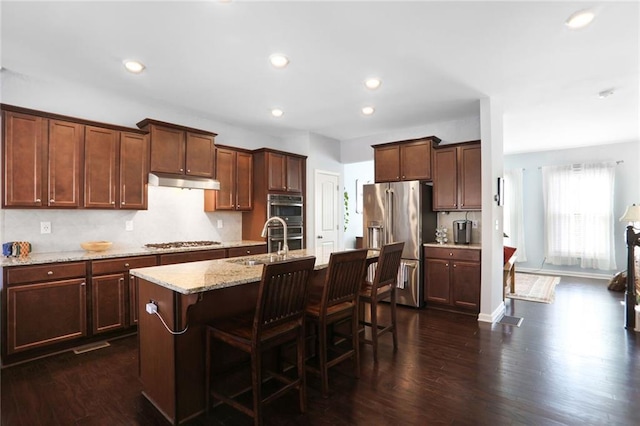 kitchen featuring a kitchen island with sink, a sink, under cabinet range hood, dark wood finished floors, and appliances with stainless steel finishes