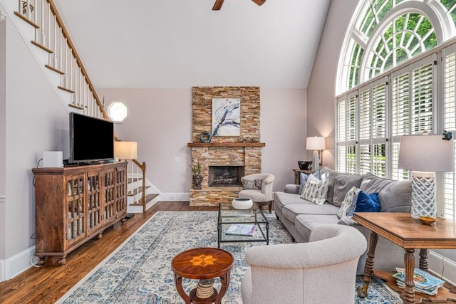 living room with stairway, ceiling fan, wood finished floors, a stone fireplace, and high vaulted ceiling