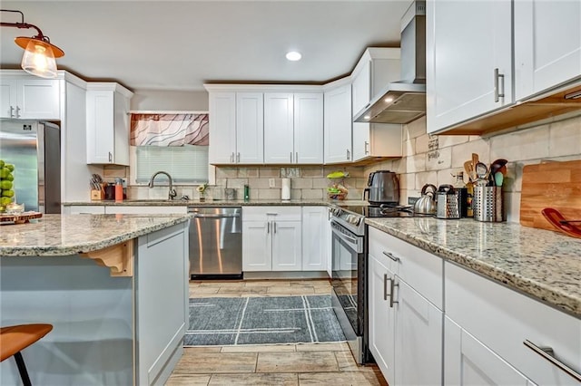 kitchen with white cabinets, wall chimney exhaust hood, appliances with stainless steel finishes, a kitchen bar, and backsplash