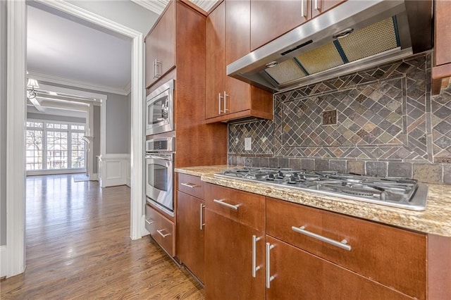 kitchen with ornamental molding, under cabinet range hood, wood finished floors, appliances with stainless steel finishes, and brown cabinetry