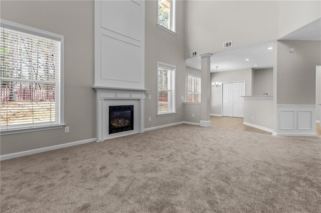 unfurnished living room featuring visible vents, a towering ceiling, a glass covered fireplace, light carpet, and ornate columns