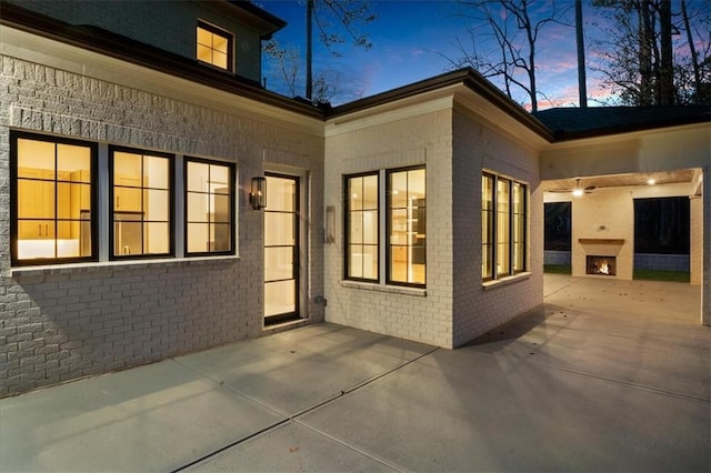 exterior space with brick siding, a patio area, a ceiling fan, and a lit fireplace