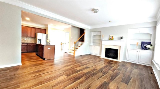 unfurnished living room featuring crown molding, a fireplace with flush hearth, built in shelves, wood finished floors, and stairway