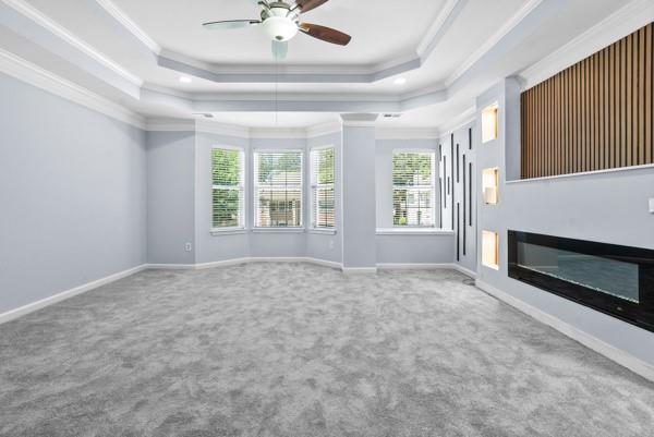unfurnished living room with a tray ceiling, carpet flooring, crown molding, a ceiling fan, and baseboards