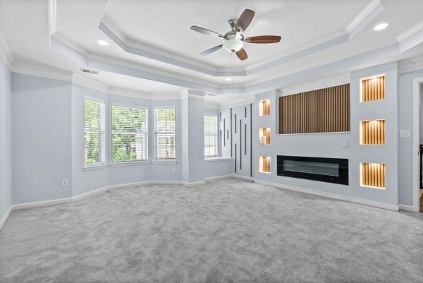 unfurnished living room with a raised ceiling, ornamental molding, carpet floors, a ceiling fan, and a glass covered fireplace