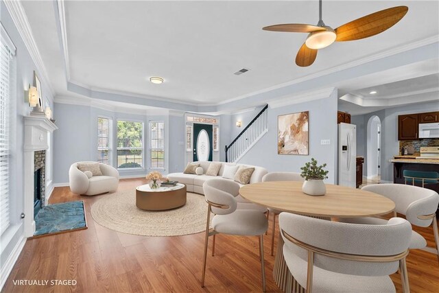 living area featuring a raised ceiling, a fireplace with flush hearth, stairs, light wood-style flooring, and a ceiling fan